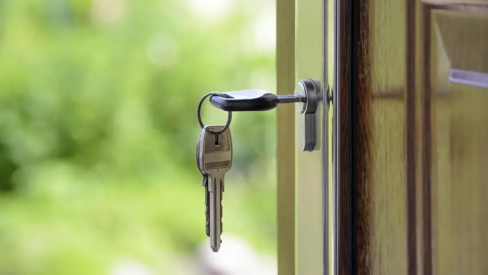 Un juego de llaves colgando de la cerradura de la puerta de una vivienda, con el jardín al fondo.